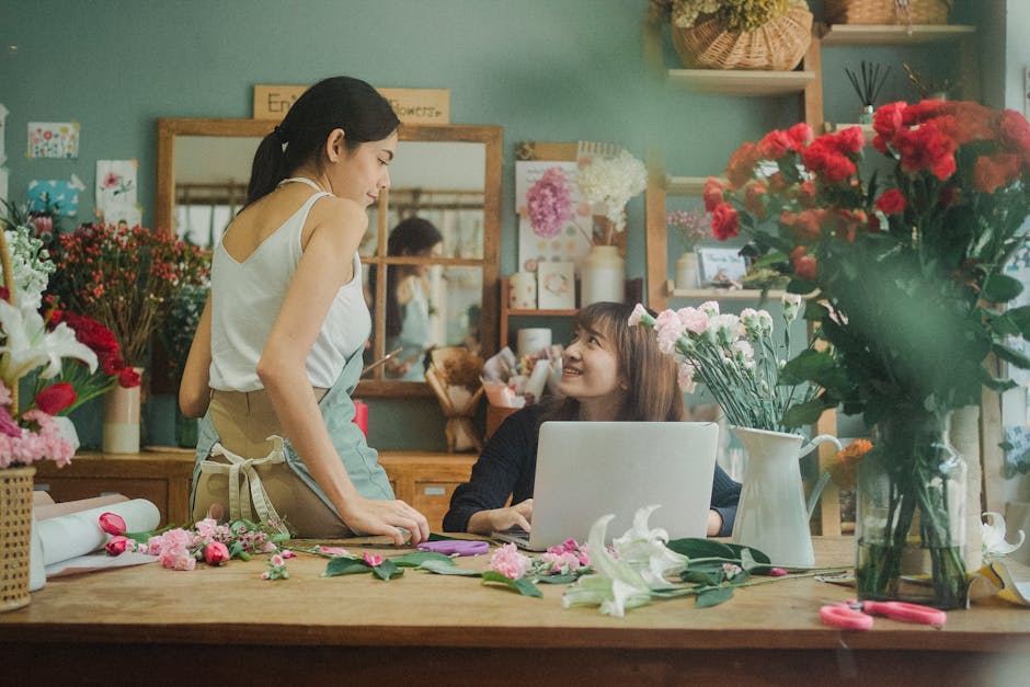 A young woman with long brown hair and a dark top is sitting at a wooden table in a flower shop, looking up and smiling at a woman with dark hair tied back, wearing a white sleeveless top and beige ap