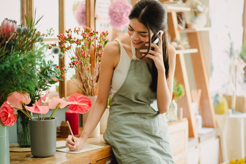 A young woman with long dark hair, smiling as she talks on a cordless phone while standing behind a wooden counter in a bright floristry shop. She is wearing a light-colored sleeveless top and a green