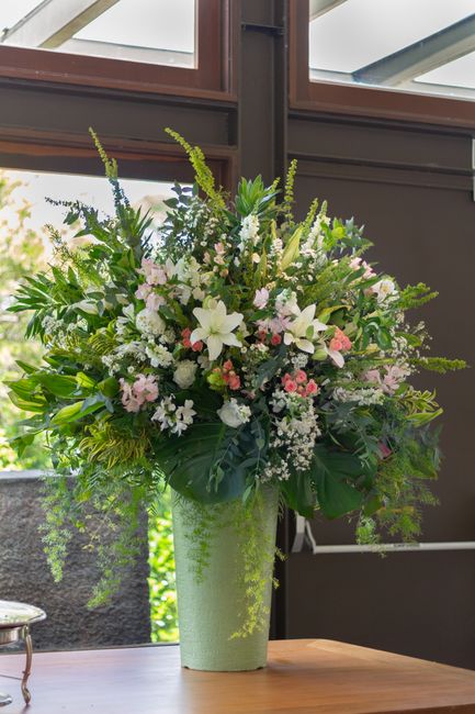 A tall, elegant floral arrangement in a light green, textured ceramic vase featuring an assortment of fresh flowers including white lilies, pink roses, white snapdragons, and delicate baby's breath, c