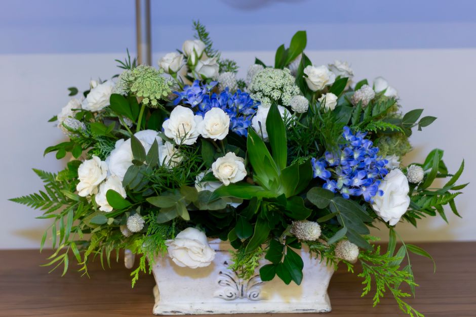 A floral arrangement featuring white roses and lisianthus, vibrant blue hydrangeas, and an assortment of greenery including ferns, eucalyptus, and other foliage, all arranged in an elegant rectangular