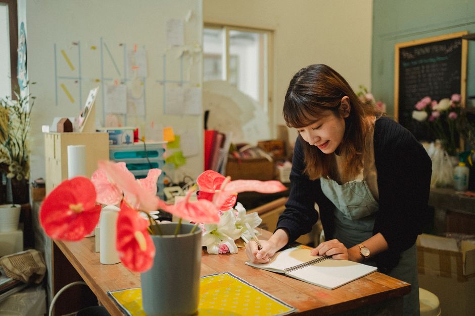 A florist with shoulder-length brown hair, wearing a beige apron over a black top, is smiling while writing in a notebook placed on a wooden work table. The table is decorated with vibrant red anthuri