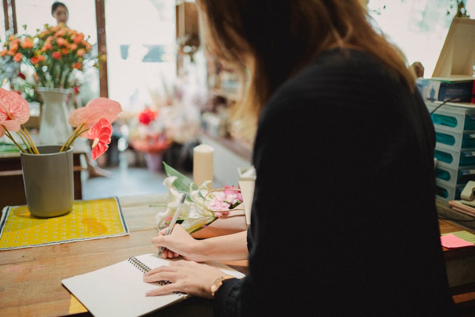 A floral arrangement being prepared at a florist shop, featuring a bouquet of pink and white flowers with green foliage, possibly roses or peonies, wrapped in soft pink paper with a white ribbon. In t