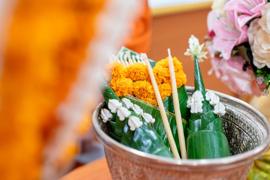 A traditional floral offering composed of bright orange marigold flowers arranged on large green banana leaves, with white jasmine flowers and slender bamboo sticks inserted into the arrangement. The