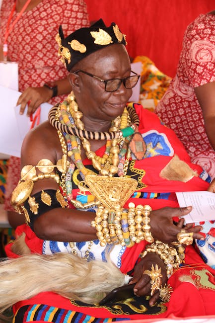 A woman dressed in vibrant, traditional African attire with intricate gold jewelry and a black headpiece decorated with gold leaves, seated in a setting with red drapes and patterned textiles. She wea