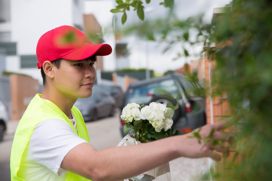 A young male flower delivery courier wearing a red cap and yellow high-visibility vest is outdoors, delivering a bouquet of fresh white hydrangeas wrapped in light-colored paper. He is reaching out wi