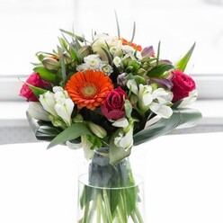 This exquisite bouquet reflects the serene elegance found around Temple's peaceful lanes and riverside gardens. Soft peach gerberas form the centrepiece-petals arranged in a mellow halo, their warm, creamy tones encircling fiery orange centres that catch the eye like lamps after dusk on the Strand. Nestled beside them, fuchsia-pink roses provide dramatic depth and a touch of opulent romance, their intricate whorls creating a sense of timeless beauty. Delicate, multi-petaled white alstroemeria and clusters of petite spray roses dot the arrangement, interspersed with lighter pink hints, diffusing a feeling of lightness and spring air. Abundant, glossy green foliage and thoughtfully curved aspidistra leaves cradle the ensemble, giving it compositional fullness and gentle containment reminiscent of Temple Garden's secluded spaces. Crystal-clear stems reveal the bouquet's freshness within a slender glass vase, while the background-a wash of soft white-illuminates every flower in tranquil, natural light. The gentle fragrance, a subtle mix of sweet rose and fresh-cut greens, lingers in the imagination. Crafted for joyful gatherings by the riverside, meaningful milestones at a local chamber, or as a sophisticated gift for someone starting a new chapter, this arrangement embodies optimism, care, and the enduring artistry of Temple's local florists. Every element is chosen to evoke a sense of calm, beauty, and new beginnings.